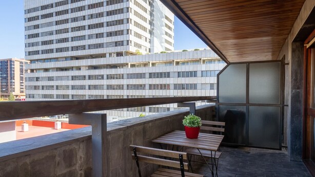Un balcon avec une table et un banc en bois, un pot de fleurs et une vue sur un immeuble moderne.