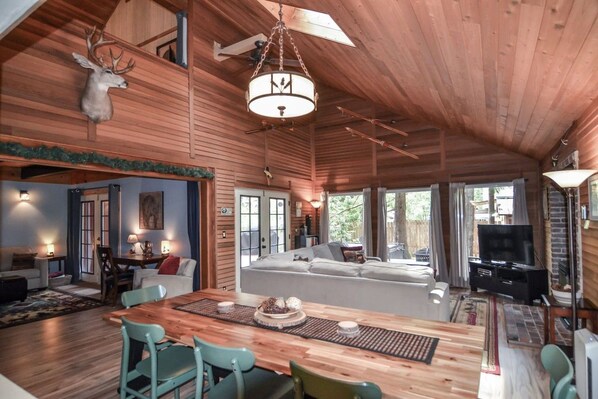 A spacious living area with a wooden ceiling, a dining table with chairs, a sofa, a television, and a fireplace.
