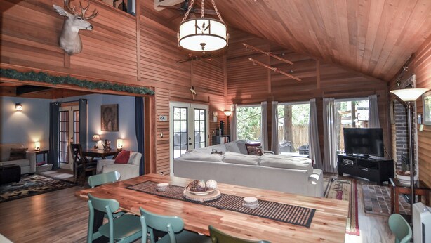 Amplia sala de estar con techo de madera, cabeza de venado montada, candelabro, sofá, televisor y una mesa de comedor con sillas.