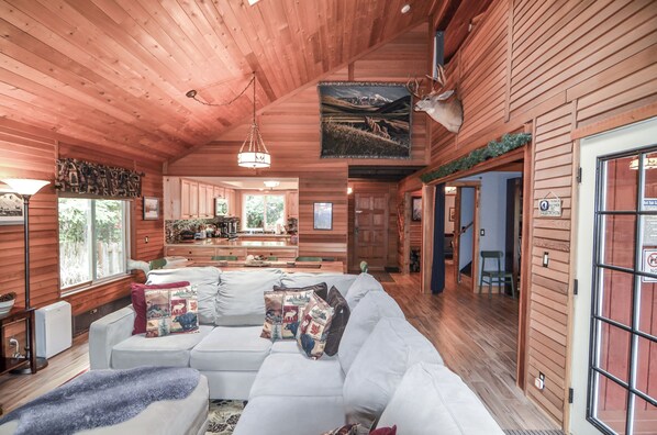 A log cabin interior with a large white sofa, wooden walls, and a dining area with a view of the outdoors.