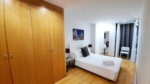 Hotellihuone, jossa on suuri puinen vaatekaappi, valkoisilla lakanoilla varustettu sänky, yöpöytä lampun kanssa ja taulu seinällä.