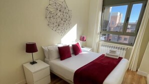 Een slaapkamer met een groot bed, wit beddengoed, rode kussens, een rode plaid en een witte nachtkastje met een lamp, plus een geometrisch wanddecoratie.