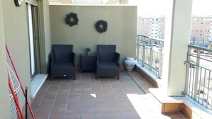 Een balkon met betegelde vloer, twee rieten stoelen, een kleine tafel en twee aan de muur bevestigde kransen.
