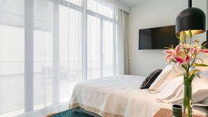 A hotel room with a large bed, a TV mounted on the wall, a vase with flowers, and a view of the outdoors through large windows.