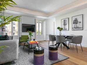 Een moderne woonkamer met een bank, salontafel en stoelen. Er is een eettafel met stoelen, een aan de muur bevestigde televisie en kunstwerken aan de wanden.