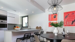 Une cuisine moderne avec des armoires blanches, un four encastré et un panier à fruits. On y trouve également un coin repas avec une table ronde, des chaises et un vase avec une plante.