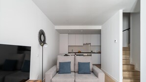 A modern living room with a grey sofa, a TV, and a floor lamp. A kitchen is visible in the background with white cabinets and a wooden staircase.