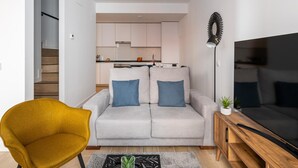 Una sala moderna con sofá, un sillón amarillo, televisión y una cocina al fondo.