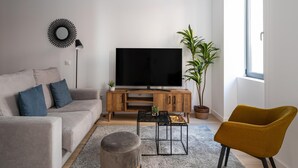 Una sala de estar moderna con un televisor de pantalla plana sobre un soporte de madera, un sofá gris con cojines azules, un sillón color mostaza, un otoman redondo pequeño, una planta en maceta y una lámpara de pie.