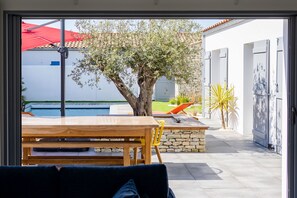 An outdoor patio with a wooden table and chairs, a stone wall, and a view of a swimming pool and a building with open doors.
