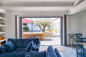 A modern living room with a sofa, a dining table, and chairs. There is a view of an outdoor patio with a table and chairs, a tree, and a building with a red roof. - La Dionysienne piscine privée (Saint-Denis-d´Oléron)