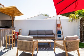 Een terras met een bank, twee rieten stoelen, een houten tafel en een rode parasol.