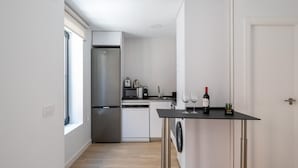 A compact kitchen with a stainless steel refrigerator, a microwave, a toaster, and a washing machine. There is a small table with two wine glasses and a bottle of wine. The room has white walls and a wooden floor.
