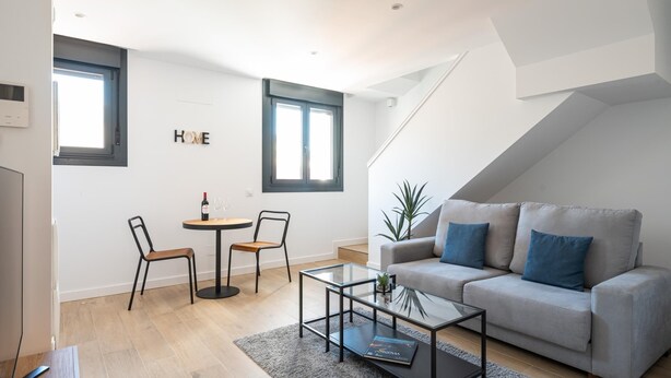 A modern living room with a sofa, a coffee table, and two chairs. There are two windows and a staircase leading to an upper level.