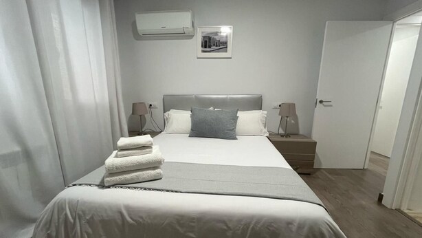 Una cama bien hecha con ropa de cama blanca, un cabecero gris y un almohadón gris. Hay dos lámparas de noche, una puerta blanca y un cuadro en la pared.