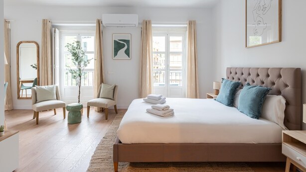 Un dormitorio con una cama grande, una zona de estar con sillas, un espejo y una ventana con cortinas.