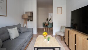 A modern living room with a grey sofa, a wooden coffee table with a vase of flowers, a flat-screen TV, and a dining area with a table and chairs. - Tirso de Molina - 1 Bedroom (Madrid)