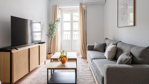 A modern living room with a flat-screen TV, a grey sofa, a wooden cabinet, and a small table with a fruit bowl. - Tirso de Molina - 1 Bedroom (Madrid)