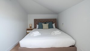 Una cama bien tendida con sábanas blancas y un cabecero marrón en una habitación pequeña y bien iluminada con paredes blancas.
