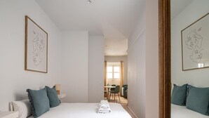 Une chambre à coucher avec un lit, des oreillers, une table de chevet et un miroir. Un couloir mène à une salle à manger équipée d’une table et de chaises.