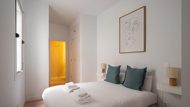 Une chambre à coucher avec un grand lit, deux lampes de chevet, une œuvre d’art encadrée au mur et une porte donnant sur une salle de bain.