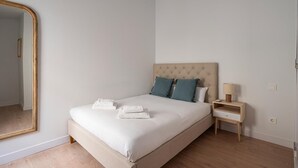 Ein schlicht eingerichtetes Schlafzimmer mit einem großen Spiegel, einem Bett mit weißen Bettwäsche und zwei Kissen, einem Nachttisch mit Lampe sowie einem Holzboden.