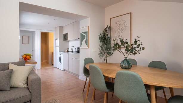 Ein modernes Wohnzimmer mit grauem Sofa, einem Holzespeisetisch mit vier Stühlen, einer Küche im Hintergrund und einem gerahmten abstrakten Gemälde an der Wand.