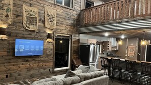 A cozy living area with a sofa, a TV mounted on the wall, and a wooden wall with hanging bags.