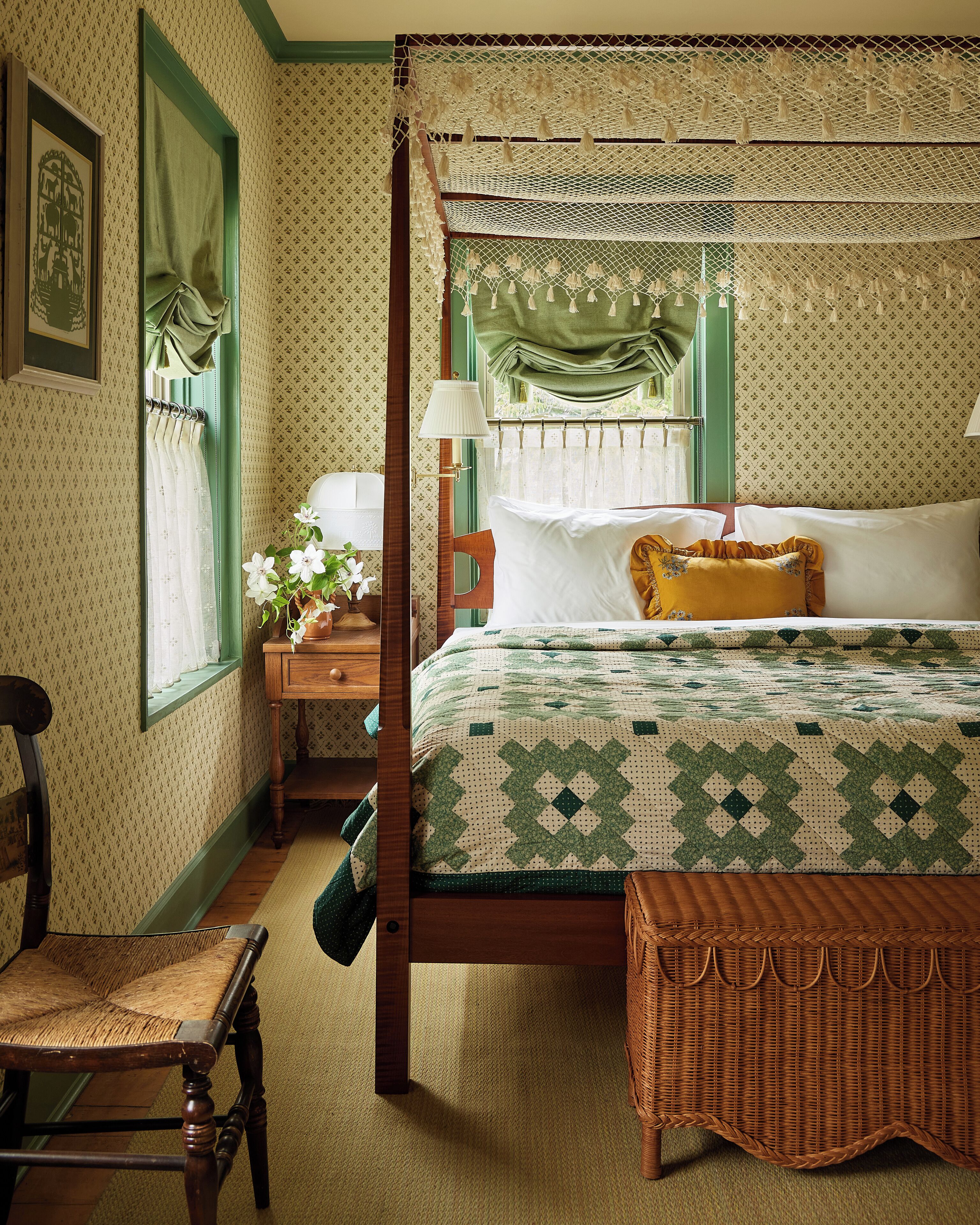 Una cama tradicional con dosel, con un edredón verde y blanco, una mesita de noche de madera con una lámpara, una silla de mimbre y un banco de madera.