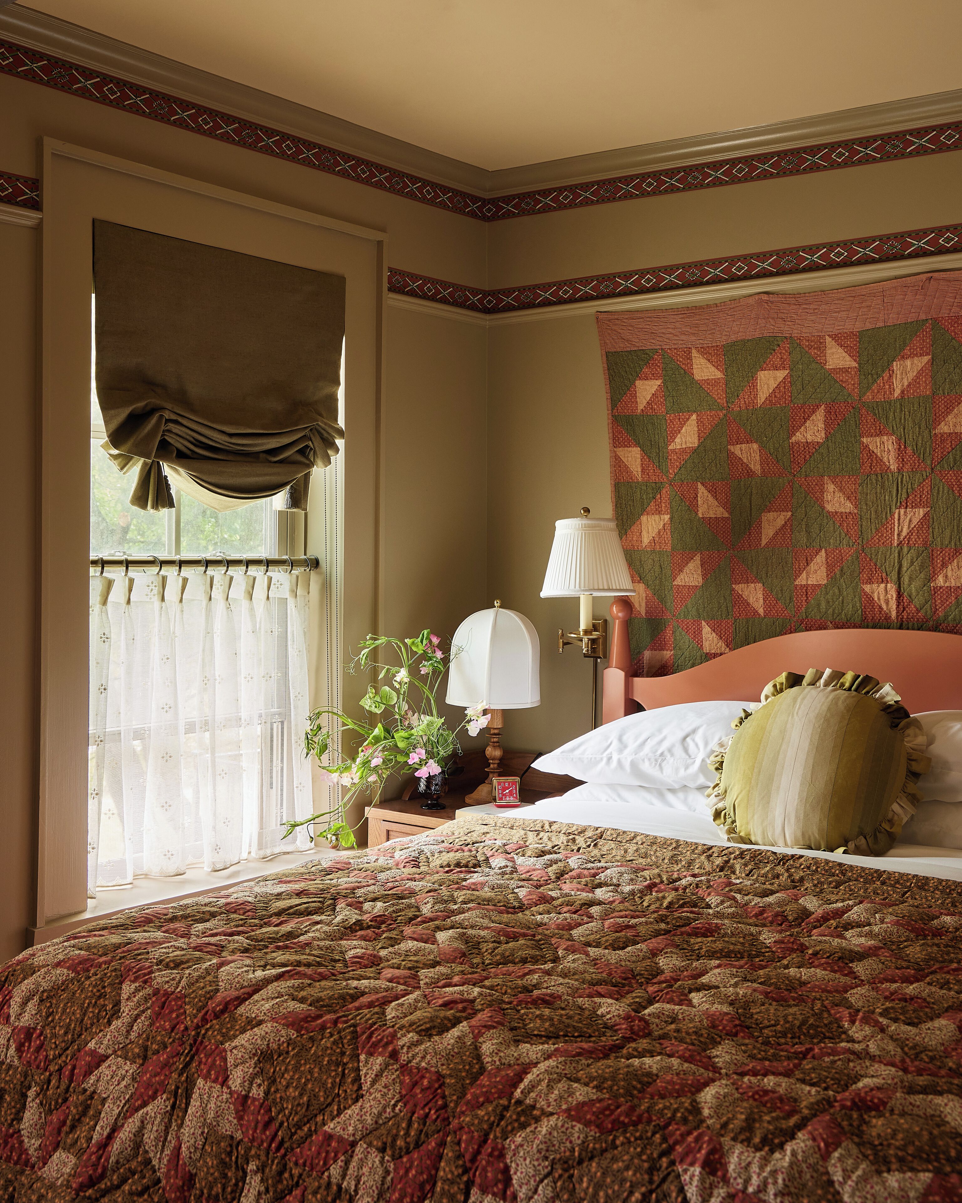 A bedroom with a large bed, a bedside table with a lamp, a window with curtains, and a patterned wall hanging.