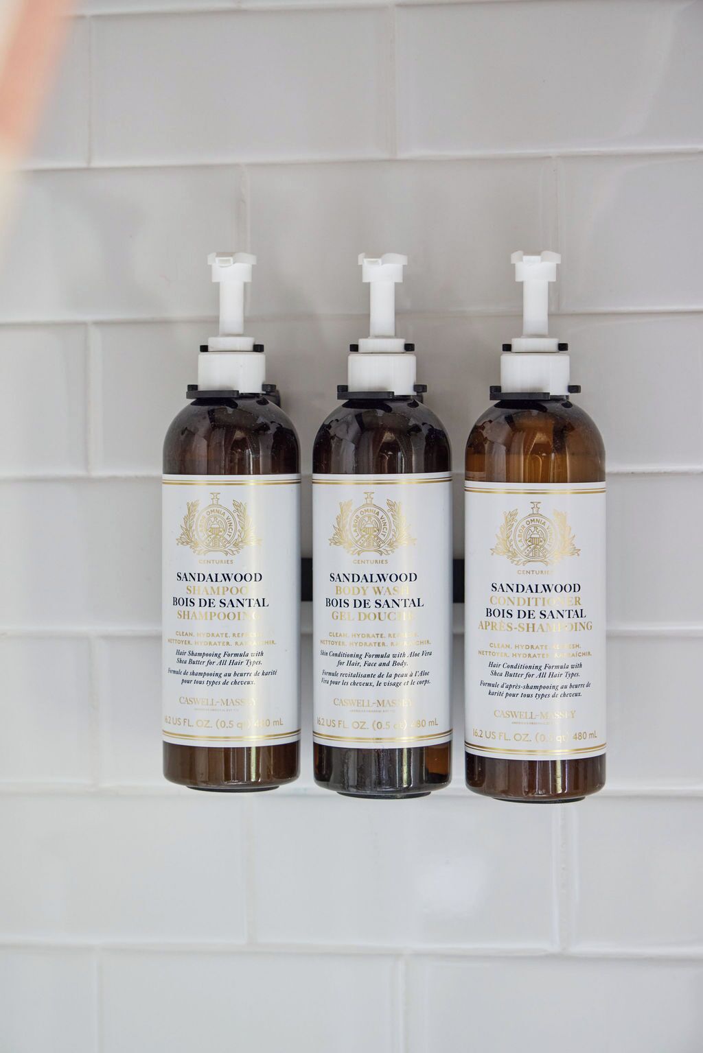 Three bottles of sandalwood body wash against a white tiled wall.