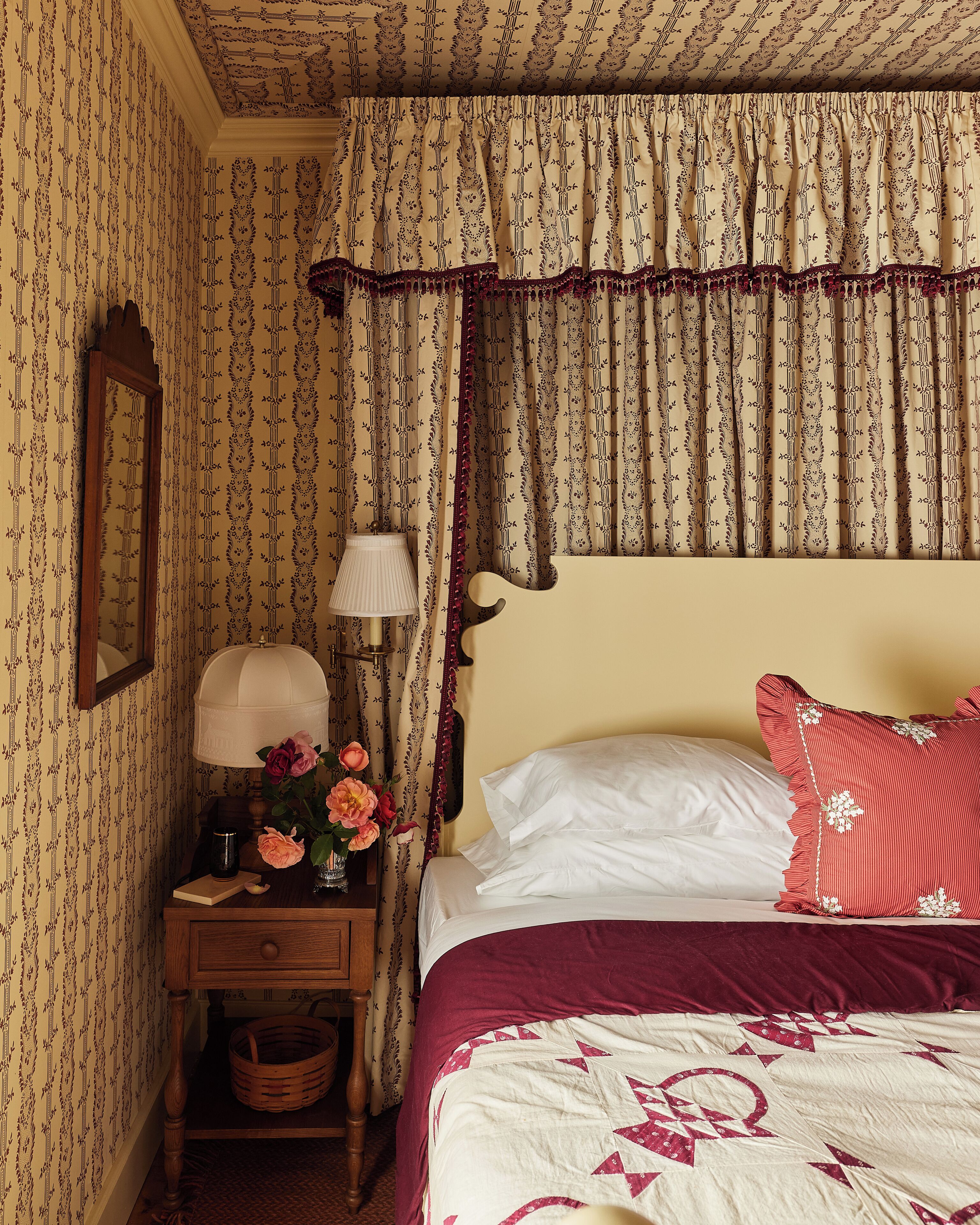 A bedroom with patterned wallpaper, a bedside table with a lamp, a vase of flowers, and a bed with a red pillow.