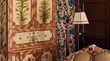 A room with a wooden cabinet with floral designs, a patterned wallpaper, a wooden door, and a quilted bedspread.