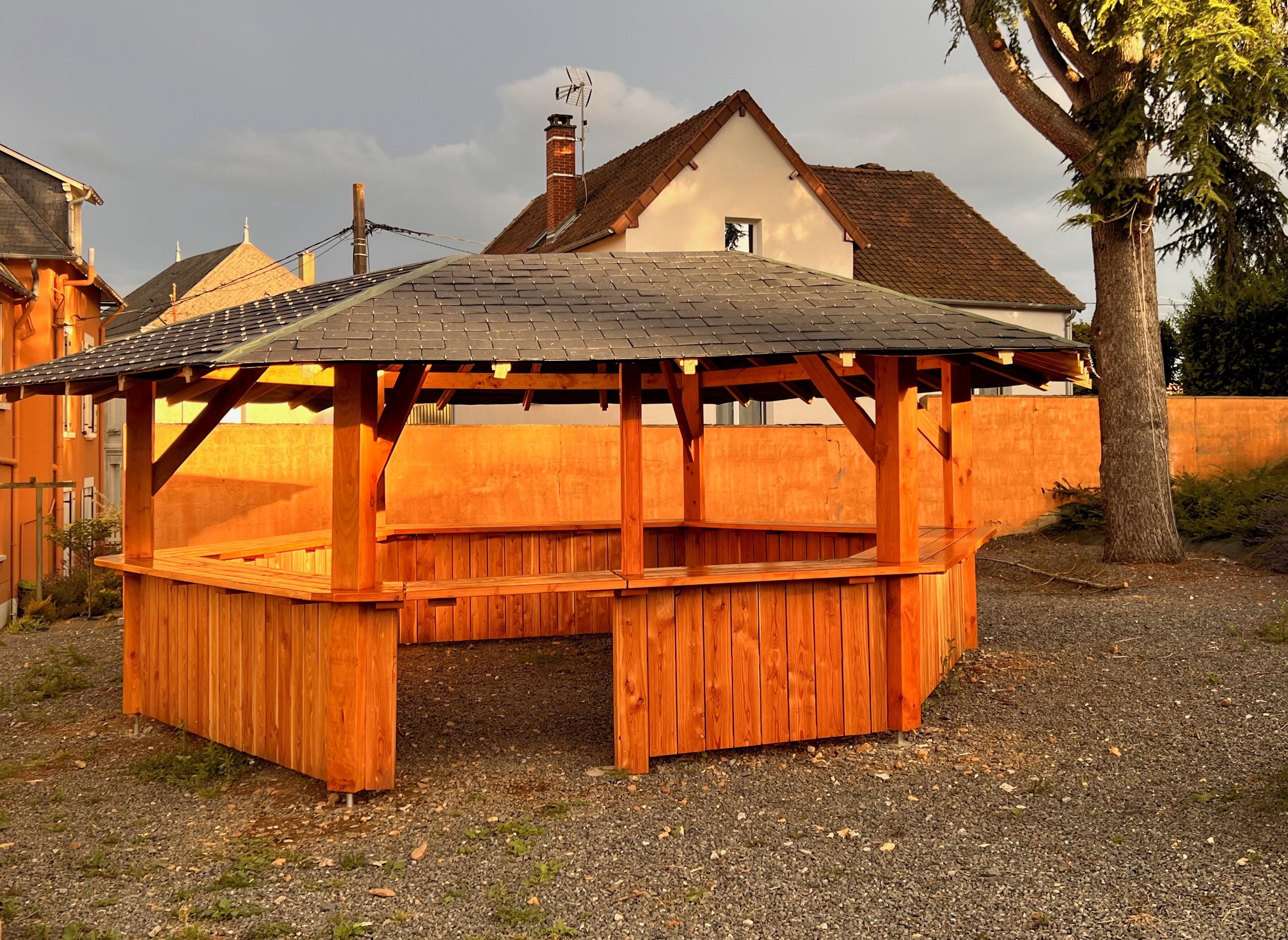 A wooden gazebo with a sloped roof, surrounded by a gravel area and a wooden fence.