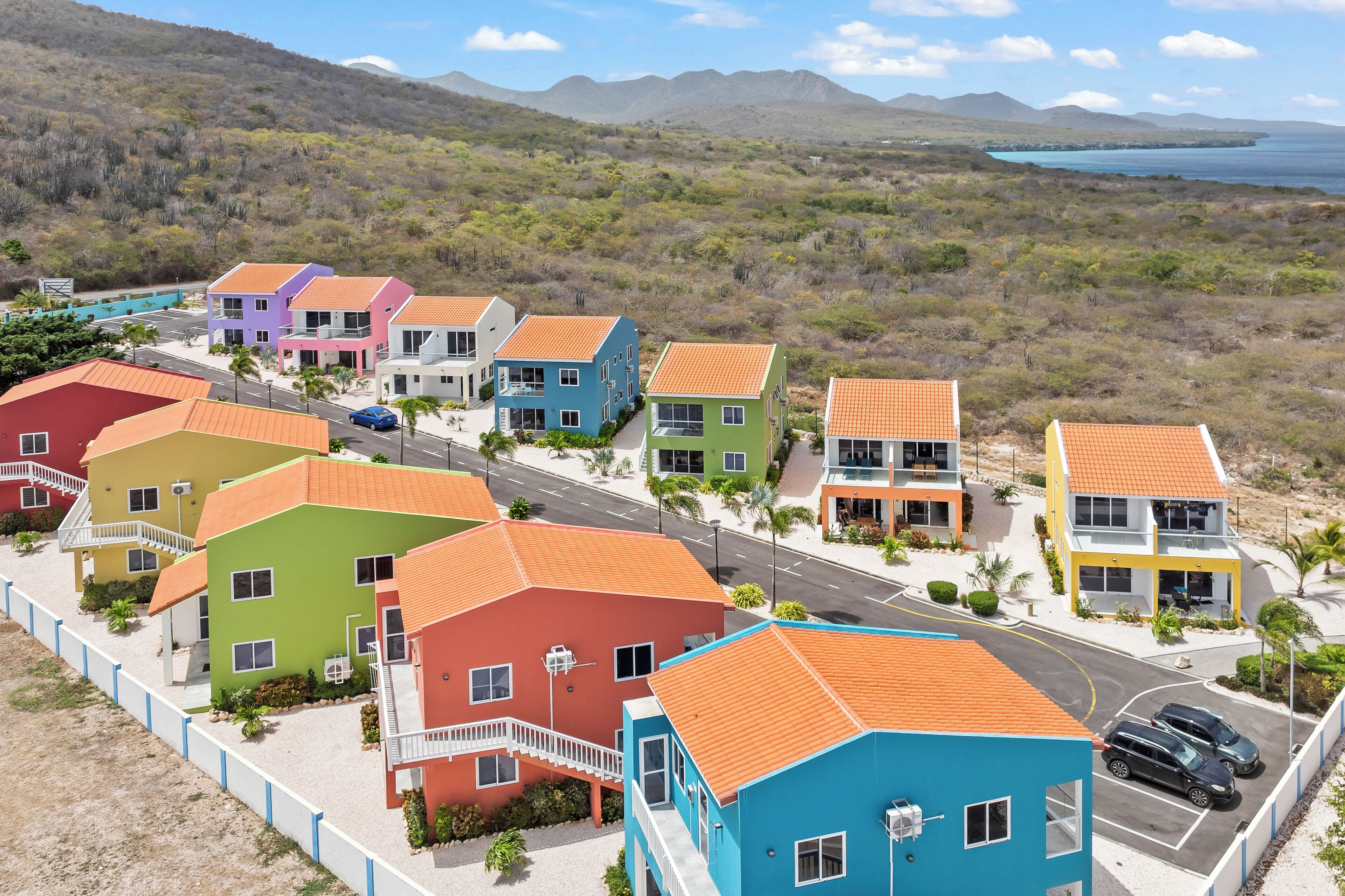 A coastal residential area with colorful houses, a road, and a view of the sea and mountains.