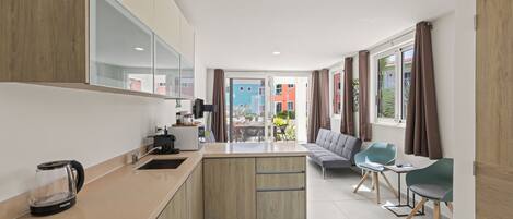 A modern kitchen with a built-in stove, a coffee maker, and a kettle on the counter. There is a dining area with a sofa and two chairs. The room has large windows with curtains.