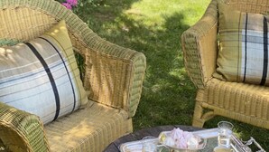 Dos sillas de mimbre con cojines a rayas, una bandeja de metal con tazas y una tetera, y un jardín con un muro de piedra y plantas.