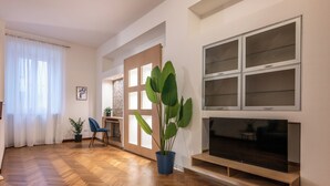 A living room with a wooden herringbone floor, a large flat-screen TV, a blue chair, and a potted plant. - Velvet Stay | Ravizza 41 by Pleiades Home (Livorno)
