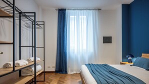 A bedroom with a bunk bed, a bed with a blue blanket, a wooden bench with towels, and a window with blue curtains. - Velvet Stay | Ravizza 41 by Pleiades Home (Livorno)
