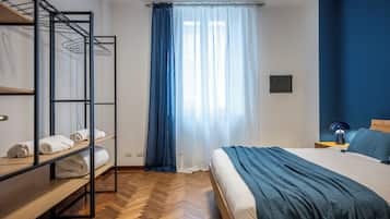 A bedroom with a bunk bed, a bed with a blue blanket, a wooden bench with towels, and a window with blue curtains.