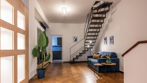 A modern interior with a herringbone wooden floor, a staircase with a metal railing, a blue sofa, and a small round table with a vase of flowers. - Velvet Stay | Ravizza 41 by Pleiades Home (Livorno)