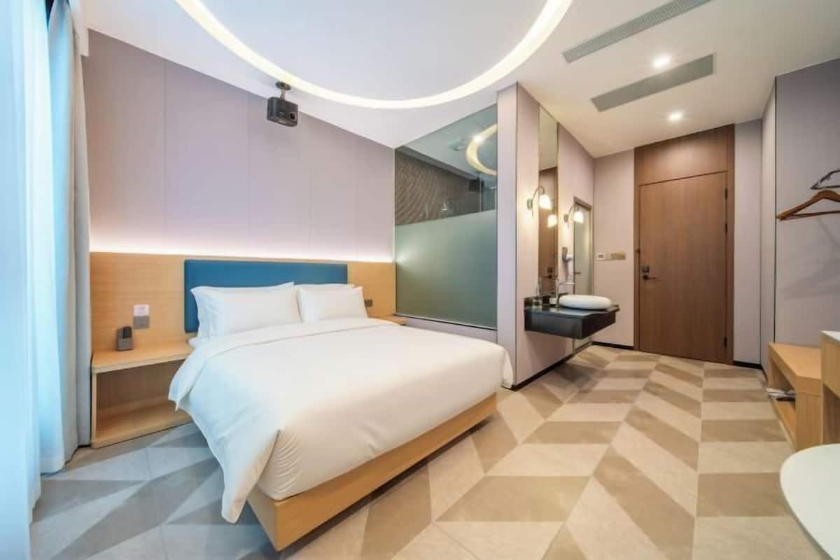 A modern hotel room with a large bed, bedside tables, a mirror, a bathroom with a sink, and a wardrobe.