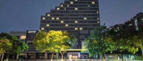 A high-rise building with illuminated windows at night, surrounded by trees and a crosswalk.