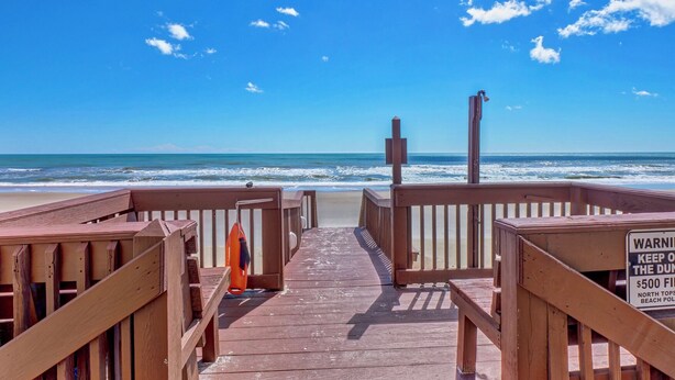Un muelle de madera que conduce a una playa con una torre de salvavidas y un letrero que advierte sobre una multa de 500 dólares por invasión de propiedad.