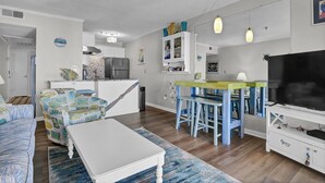 Un espacio habitable compacto con kitchenette, una zona de comedor con sillas azules, un sofá blanco y un televisor sobre un mueble blanco.