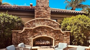 Un patio exterior con mobiliario de mimbre, una chimenea de piedra y vistas a una casa con techo de tejas.