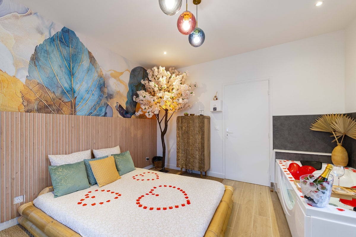 A bedroom with a bed, pillows, a wall mural, a decorative tree, and a bedside table with a bottle and glasses.