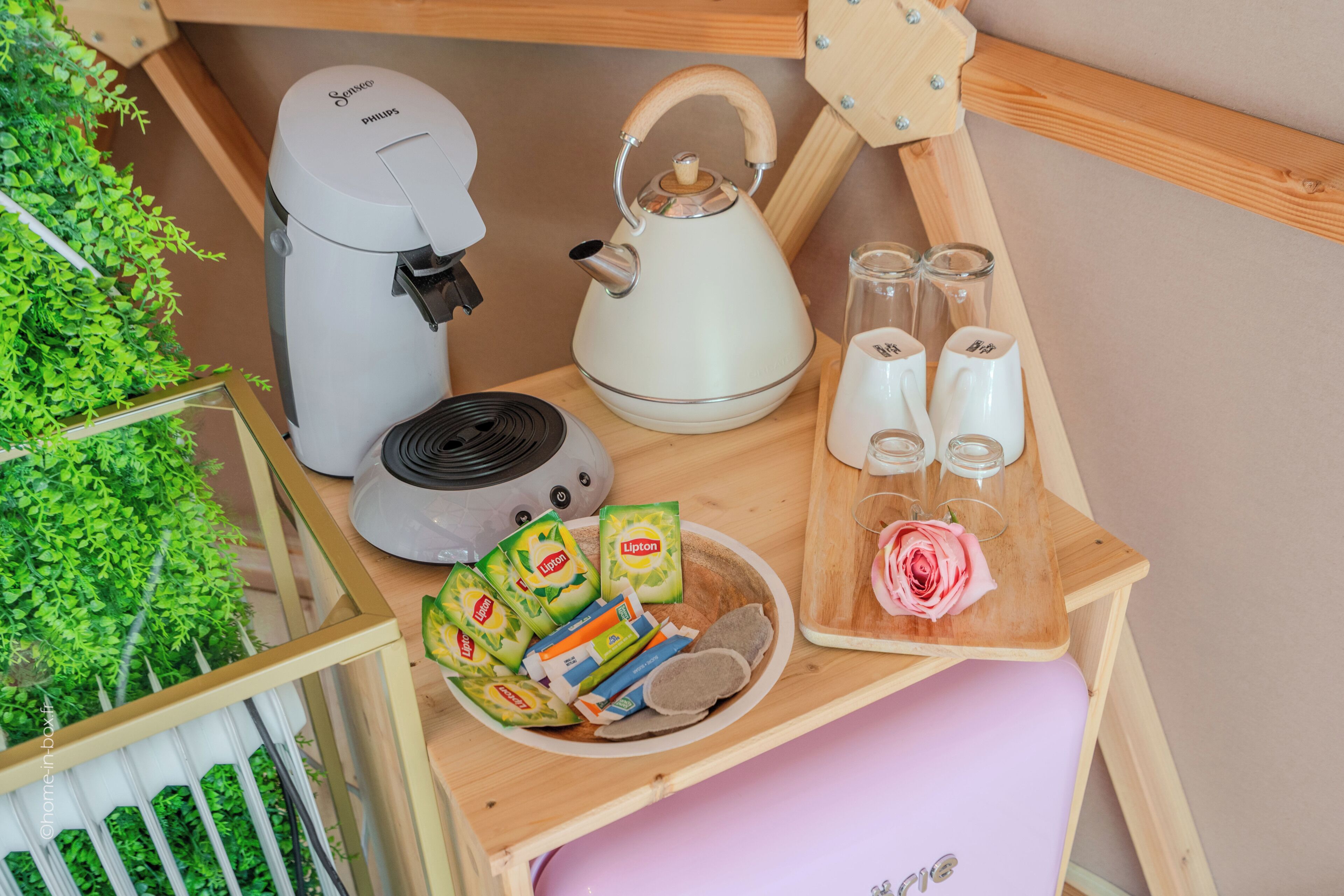 Et tekøkken med kaffemaskine, elkedel og tesæt, samt emballerede madvarer og en skærebræt med en rose.