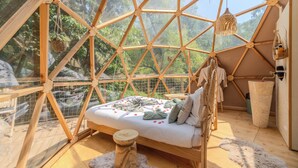 Habitación en forma de cúpula geométrica con cama, piso de madera y vista a los árboles a través de sus paredes transparentes.