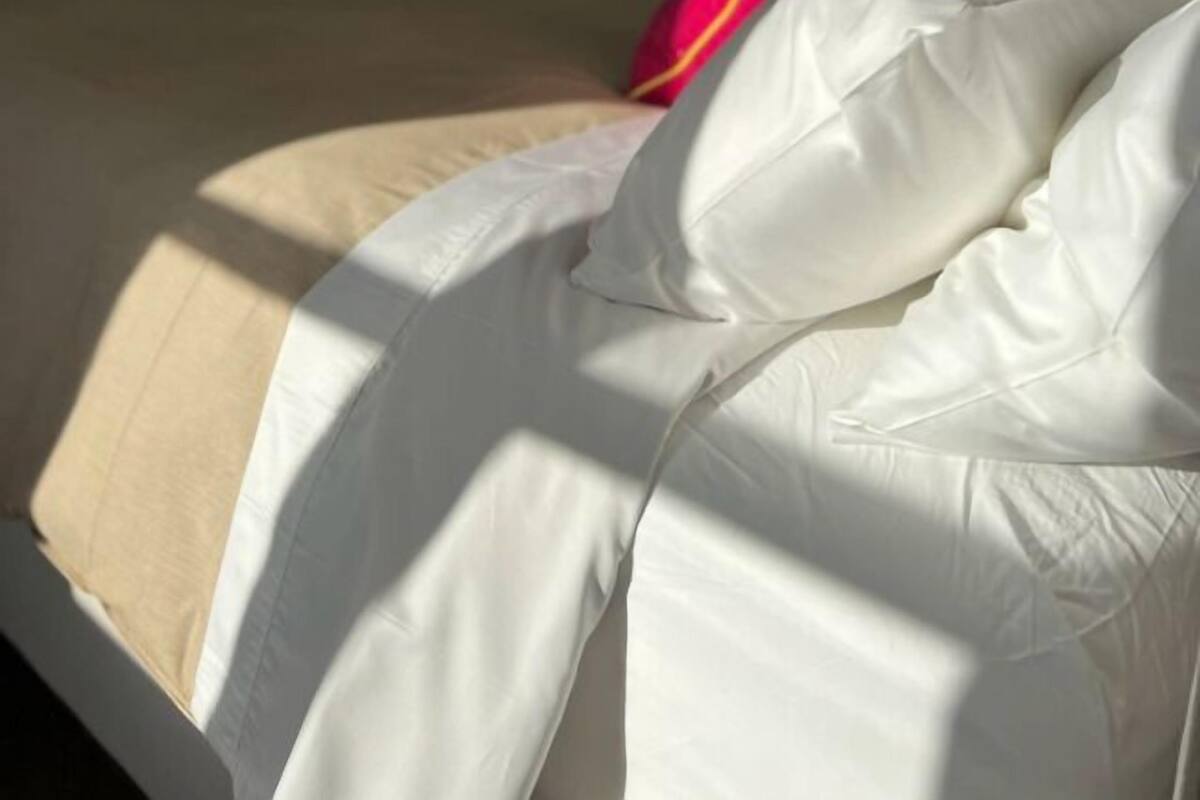 A bed with white bedding and a beige pillow, a red and yellow blanket, and a painting on the wall.